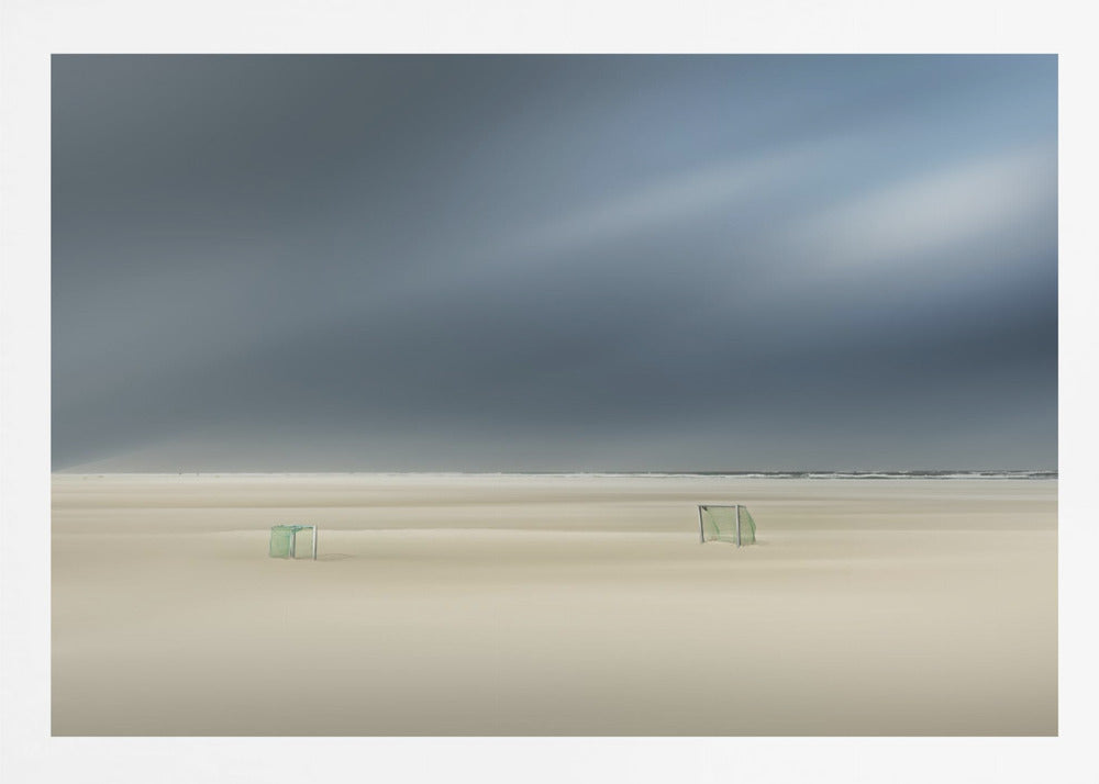 A minimalist long-exposure photograph of a vast, empty beach under a dark, moody sky. Two small, solitary soccer goals sit on the pale sand, with the ocean visible as a thin line in the distance. The artwork is presented in a silver frame. Artwork
