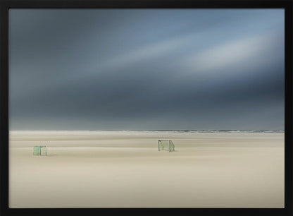 A minimalist long-exposure photograph of a vast, empty beach under a dark, moody sky. Two small, solitary soccer goals sit on the pale sand, with the ocean visible as a thin line in the distance. The artwork is presented in a silver frame. Artwork