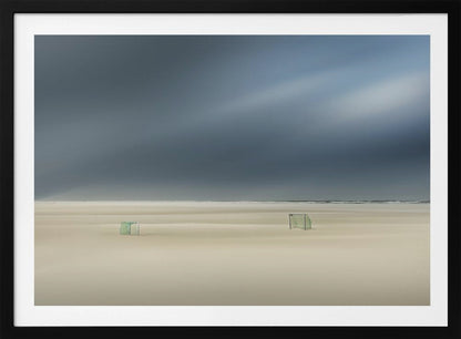A minimalist long-exposure photograph of a vast, empty beach under a dark, moody sky. Two small, solitary soccer goals sit on the pale sand, with the ocean visible as a thin line in the distance. The artwork is presented in a silver frame. Artwork