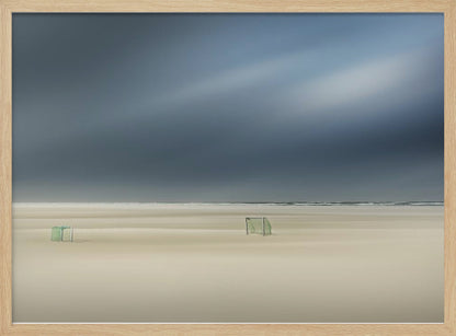 A minimalist long-exposure photograph of a vast, empty beach under a dark, moody sky. Two small, solitary soccer goals sit on the pale sand, with the ocean visible as a thin line in the distance. The artwork is presented in a silver frame. Artwork