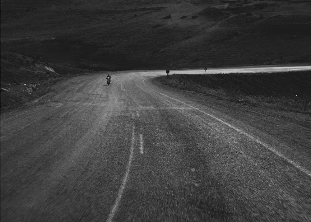 A moody, black and white photograph capturing a lone motorcyclist from behind as they navigate a curving road through a dark, hilly terrain, framed in silver. Decor
