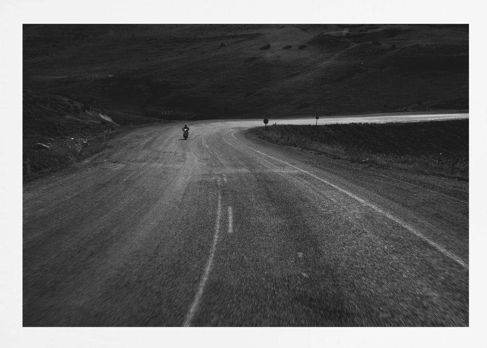 A moody, black and white photograph capturing a lone motorcyclist from behind as they navigate a curving road through a dark, hilly terrain, framed in silver. Decor