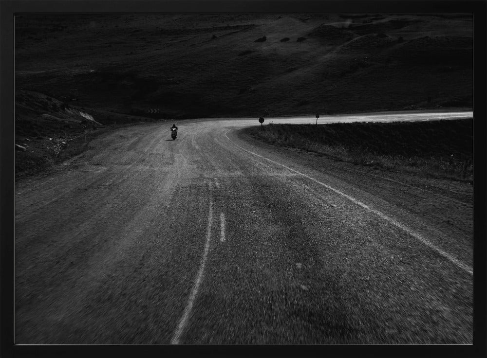 A moody, black and white photograph capturing a lone motorcyclist from behind as they navigate a curving road through a dark, hilly terrain, framed in silver. Decor