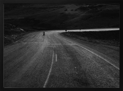 A moody, black and white photograph capturing a lone motorcyclist from behind as they navigate a curving road through a dark, hilly terrain, framed in silver. Decor