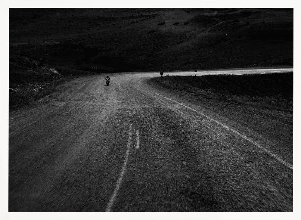 A moody, black and white photograph capturing a lone motorcyclist from behind as they navigate a curving road through a dark, hilly terrain, framed in silver. Decor