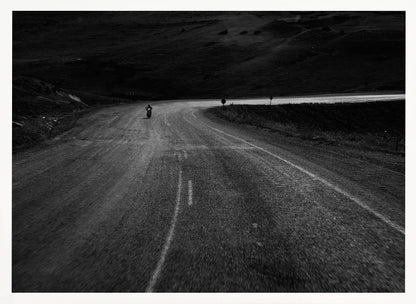 A moody, black and white photograph capturing a lone motorcyclist from behind as they navigate a curving road through a dark, hilly terrain, framed in silver. Decor