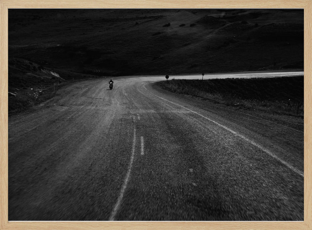 A moody, black and white photograph capturing a lone motorcyclist from behind as they navigate a curving road through a dark, hilly terrain, framed in silver. Decor