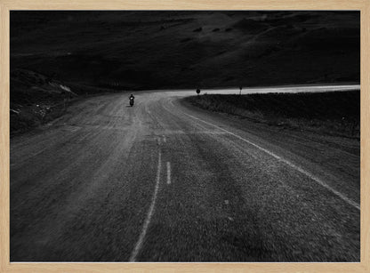 A moody, black and white photograph capturing a lone motorcyclist from behind as they navigate a curving road through a dark, hilly terrain, framed in silver. Decor