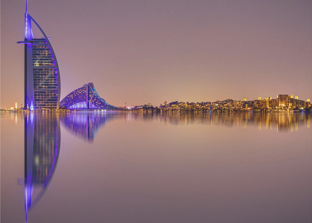 A framed panoramic night photograph of the Dubai skyline featuring the Burj Al Arab hotel lit up in brilliant purple. The city lights and architecture are perfectly reflected in the calm water in the foreground, all under a hazy, dusky sky. Poster