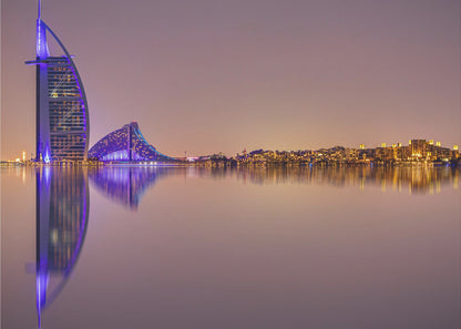 A framed panoramic night photograph of the Dubai skyline featuring the Burj Al Arab hotel lit up in brilliant purple. The city lights and architecture are perfectly reflected in the calm water in the foreground, all under a hazy, dusky sky. Poster