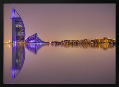 A framed panoramic night photograph of the Dubai skyline featuring the Burj Al Arab hotel lit up in brilliant purple. The city lights and architecture are perfectly reflected in the calm water in the foreground, all under a hazy, dusky sky. Poster