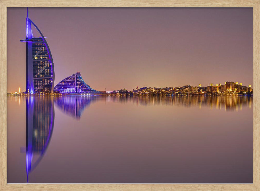 A framed panoramic night photograph of the Dubai skyline featuring the Burj Al Arab hotel lit up in brilliant purple. The city lights and architecture are perfectly reflected in the calm water in the foreground, all under a hazy, dusky sky. Poster