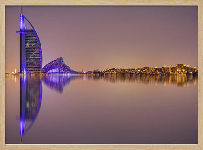 A framed panoramic night photograph of the Dubai skyline featuring the Burj Al Arab hotel lit up in brilliant purple. The city lights and architecture are perfectly reflected in the calm water in the foreground, all under a hazy, dusky sky. Poster