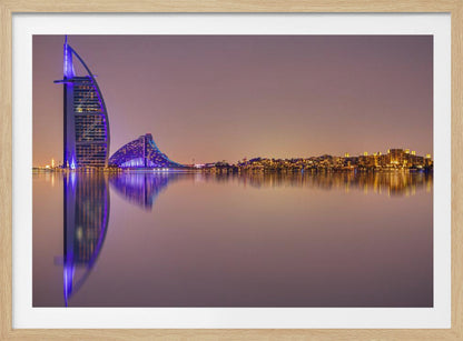 A framed panoramic night photograph of the Dubai skyline featuring the Burj Al Arab hotel lit up in brilliant purple. The city lights and architecture are perfectly reflected in the calm water in the foreground, all under a hazy, dusky sky. Poster