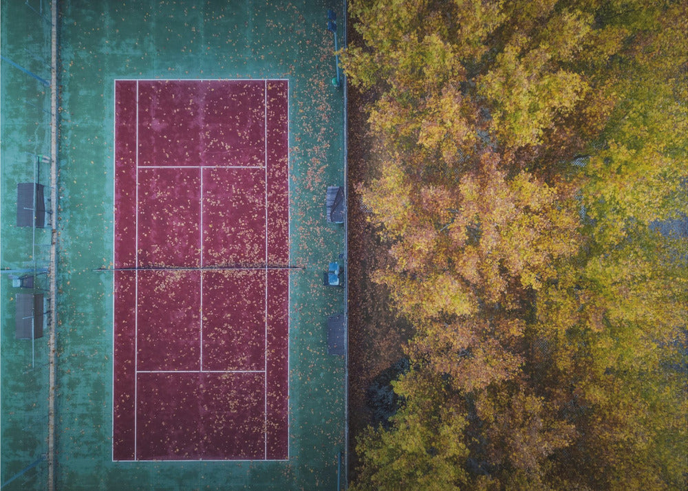 An aerial top-down photograph of a red and green tennis court covered in fallen leaves, adjacent to a dense grove of trees with vibrant yellow and gold autumn foliage, all enclosed in a silver frame. Wall Art