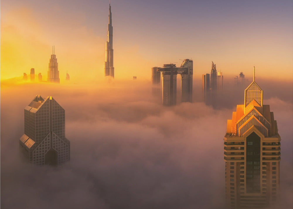 An aerial photograph of the Dubai skyline shrouded in a thick blanket of fog at sunrise. The warm golden light illuminates the tops of several skyscrapers, including the Burj Khalifa, which pierce through the clouds creating a dreamlike and majestic scene. Print