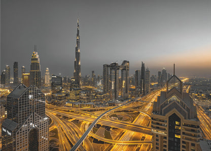 A framed, high-angle, long-exposure photograph of the Dubai skyline at dusk. The towering Burj Khalifa stands out against a grey sky, while the city's skyscrapers are illuminated. In the foreground, a complex network of highways glows with the yellow light trails of traffic, creating intricate patterns. Poster