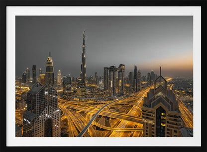 A framed, high-angle, long-exposure photograph of the Dubai skyline at dusk. The towering Burj Khalifa stands out against a grey sky, while the city's skyscrapers are illuminated. In the foreground, a complex network of highways glows with the yellow light trails of traffic, creating intricate patterns. Poster