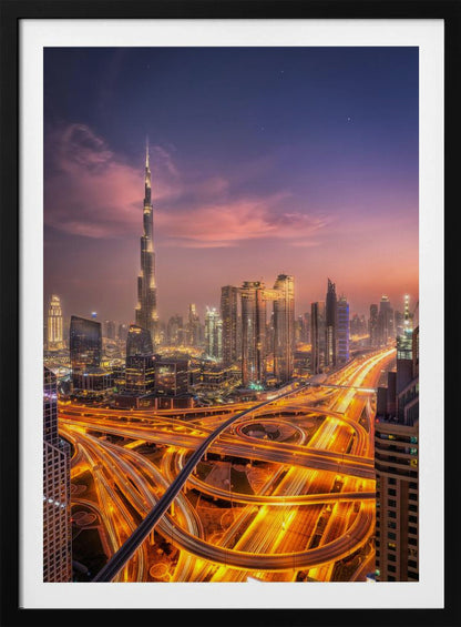 A stunning aerial photograph of the Dubai skyline at dusk, featuring the illuminated Burj Khalifa against a purple and pink sunset sky. In the foreground, complex highway interchanges are aglow with the golden-orange light trails of moving traffic, creating a sense of vibrant energy. Poster