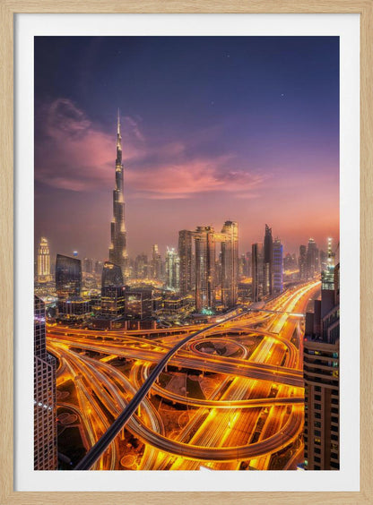 A stunning aerial photograph of the Dubai skyline at dusk, featuring the illuminated Burj Khalifa against a purple and pink sunset sky. In the foreground, complex highway interchanges are aglow with the golden-orange light trails of moving traffic, creating a sense of vibrant energy. Poster