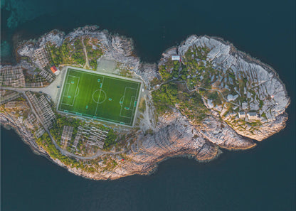An aerial view of a vibrant green soccer field situated on a small, rocky island surrounded by dark blue water. The photograph, taken from directly above, captures the unique and remote location of the sports facility, with small buildings and wooden structures dotting the island's terrain. The entire scene is enclosed within a silver frame. Print