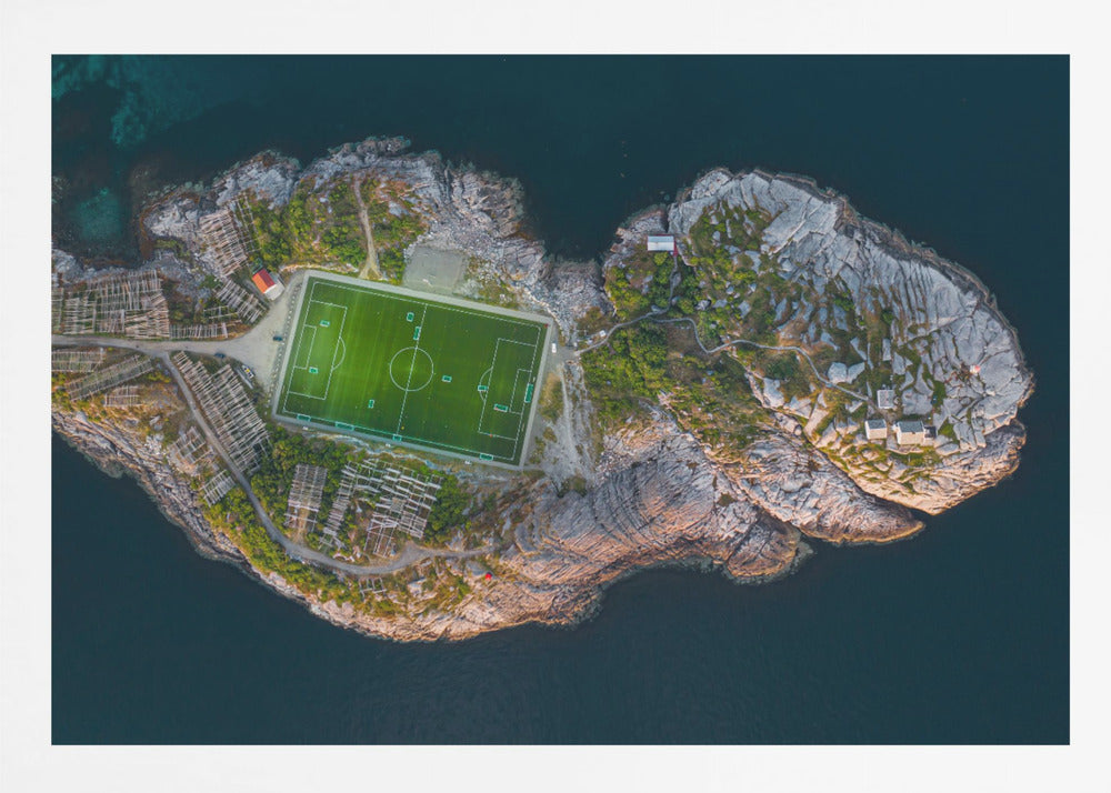 An aerial view of a vibrant green soccer field situated on a small, rocky island surrounded by dark blue water. The photograph, taken from directly above, captures the unique and remote location of the sports facility, with small buildings and wooden structures dotting the island's terrain. The entire scene is enclosed within a silver frame. Print
