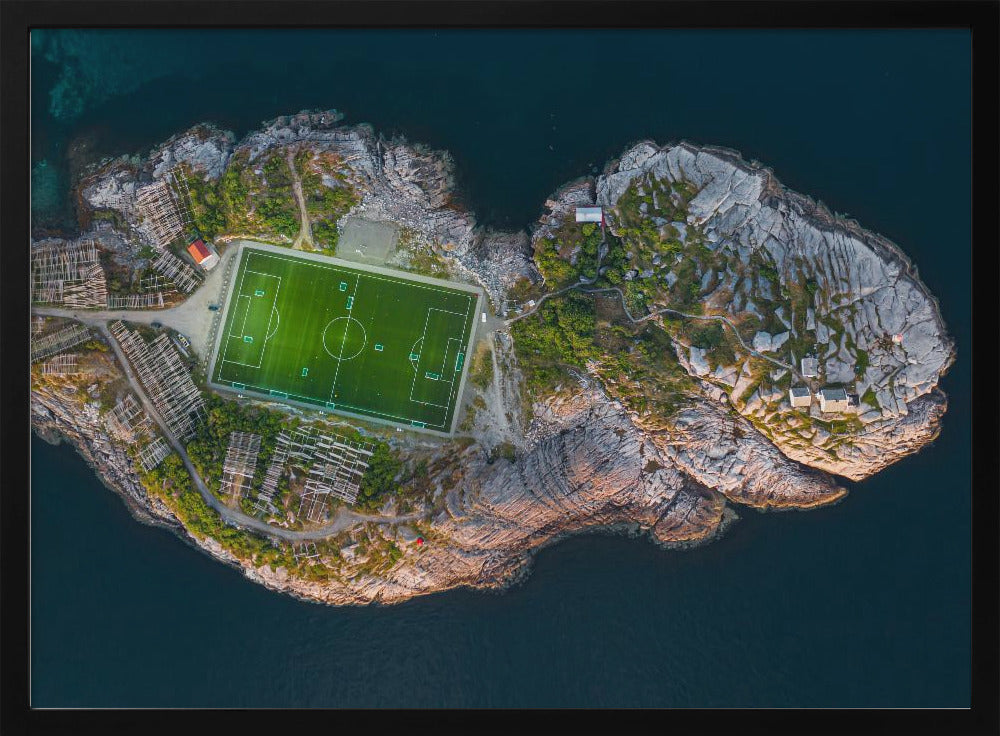 An aerial view of a vibrant green soccer field situated on a small, rocky island surrounded by dark blue water. The photograph, taken from directly above, captures the unique and remote location of the sports facility, with small buildings and wooden structures dotting the island's terrain. The entire scene is enclosed within a silver frame. Print