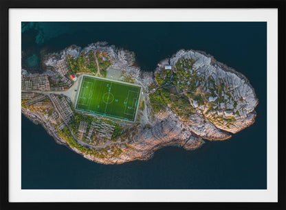 An aerial view of a vibrant green soccer field situated on a small, rocky island surrounded by dark blue water. The photograph, taken from directly above, captures the unique and remote location of the sports facility, with small buildings and wooden structures dotting the island's terrain. The entire scene is enclosed within a silver frame. Print
