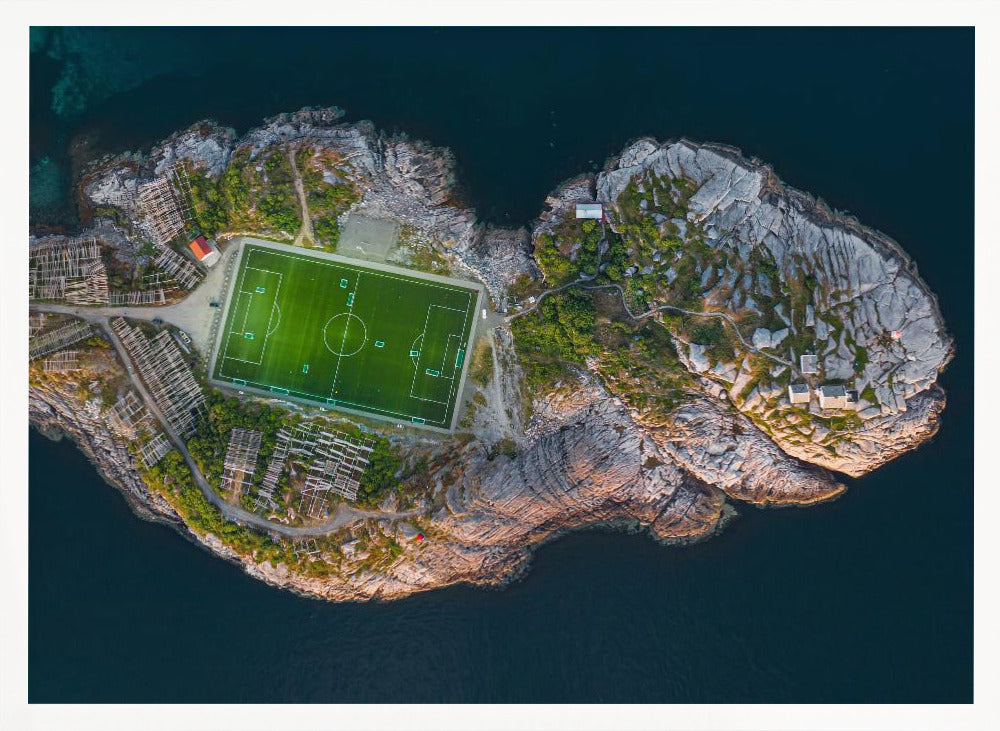 An aerial view of a vibrant green soccer field situated on a small, rocky island surrounded by dark blue water. The photograph, taken from directly above, captures the unique and remote location of the sports facility, with small buildings and wooden structures dotting the island's terrain. The entire scene is enclosed within a silver frame. Print