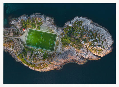 An aerial view of a vibrant green soccer field situated on a small, rocky island surrounded by dark blue water. The photograph, taken from directly above, captures the unique and remote location of the sports facility, with small buildings and wooden structures dotting the island's terrain. The entire scene is enclosed within a silver frame. Print