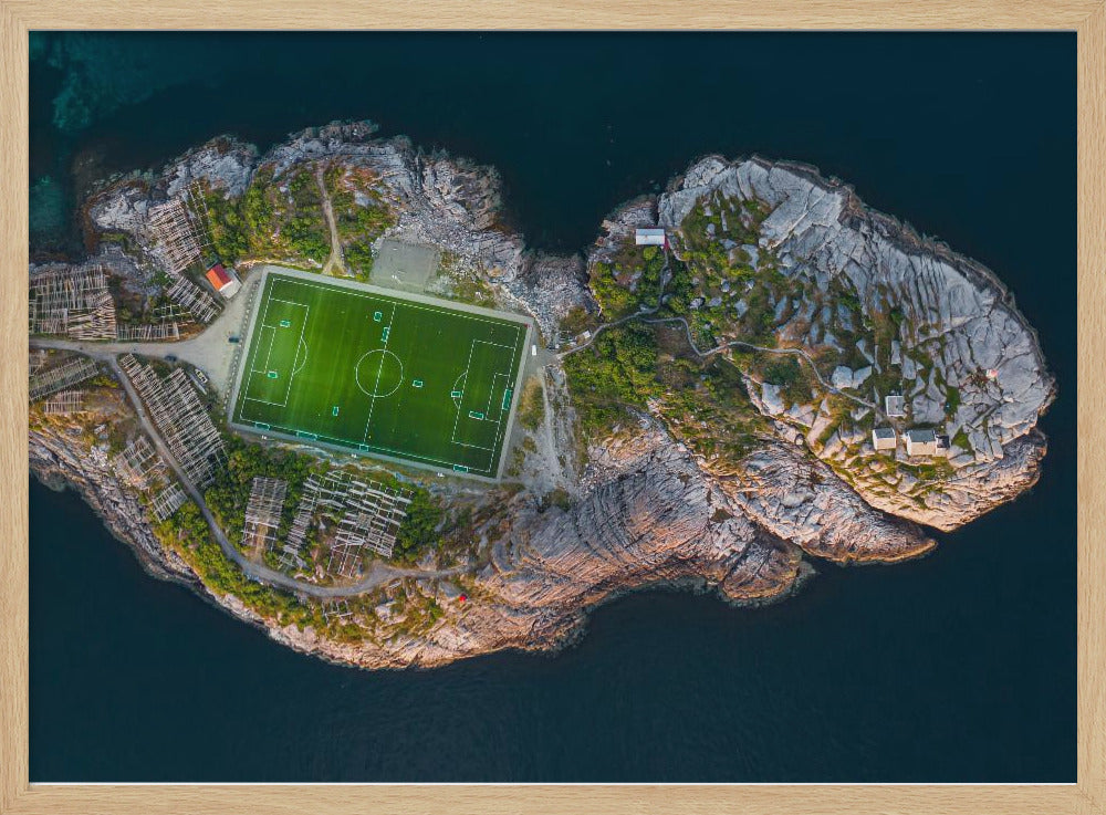 An aerial view of a vibrant green soccer field situated on a small, rocky island surrounded by dark blue water. The photograph, taken from directly above, captures the unique and remote location of the sports facility, with small buildings and wooden structures dotting the island's terrain. The entire scene is enclosed within a silver frame. Print