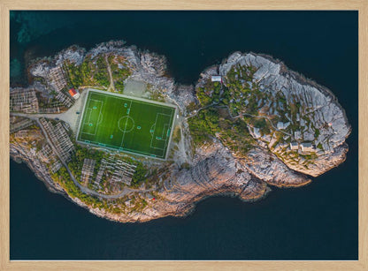 An aerial view of a vibrant green soccer field situated on a small, rocky island surrounded by dark blue water. The photograph, taken from directly above, captures the unique and remote location of the sports facility, with small buildings and wooden structures dotting the island's terrain. The entire scene is enclosed within a silver frame. Print