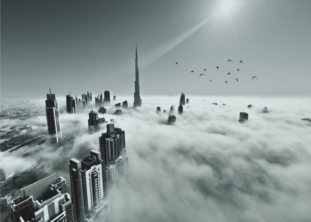 A framed, black and white aerial photograph of the Dubai skyline piercing through a thick layer of clouds. The Burj Khalifa stands prominently in the center, with other skyscrapers emerging from the fog. A flock of birds flies in the sky as a bright sunbeam shines down from the upper right corner. Wall Art