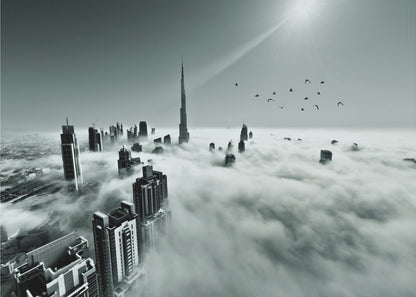 A framed, black and white aerial photograph of the Dubai skyline piercing through a thick layer of clouds. The Burj Khalifa stands prominently in the center, with other skyscrapers emerging from the fog. A flock of birds flies in the sky as a bright sunbeam shines down from the upper right corner. Wall Art