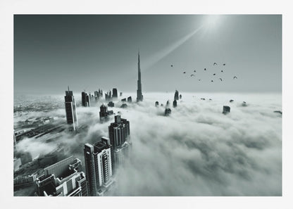 A framed, black and white aerial photograph of the Dubai skyline piercing through a thick layer of clouds. The Burj Khalifa stands prominently in the center, with other skyscrapers emerging from the fog. A flock of birds flies in the sky as a bright sunbeam shines down from the upper right corner. Wall Art