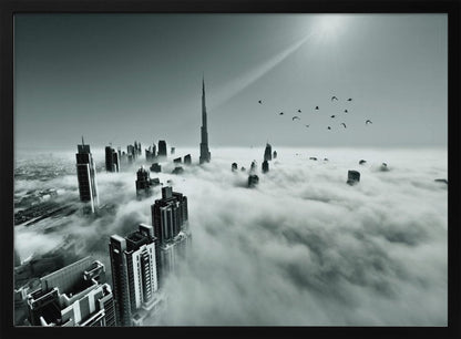 A framed, black and white aerial photograph of the Dubai skyline piercing through a thick layer of clouds. The Burj Khalifa stands prominently in the center, with other skyscrapers emerging from the fog. A flock of birds flies in the sky as a bright sunbeam shines down from the upper right corner. Wall Art