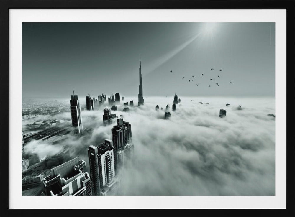 A framed, black and white aerial photograph of the Dubai skyline piercing through a thick layer of clouds. The Burj Khalifa stands prominently in the center, with other skyscrapers emerging from the fog. A flock of birds flies in the sky as a bright sunbeam shines down from the upper right corner. Wall Art
