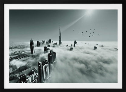 A framed, black and white aerial photograph of the Dubai skyline piercing through a thick layer of clouds. The Burj Khalifa stands prominently in the center, with other skyscrapers emerging from the fog. A flock of birds flies in the sky as a bright sunbeam shines down from the upper right corner. Wall Art