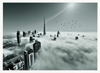A framed, black and white aerial photograph of the Dubai skyline piercing through a thick layer of clouds. The Burj Khalifa stands prominently in the center, with other skyscrapers emerging from the fog. A flock of birds flies in the sky as a bright sunbeam shines down from the upper right corner. Wall Art