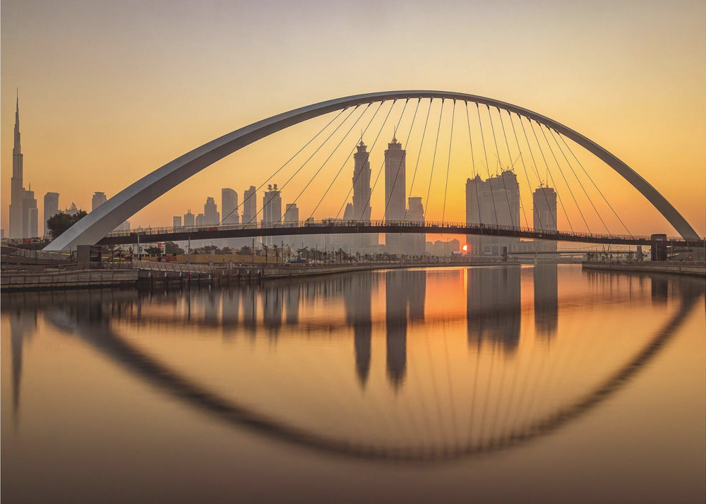 A panoramic photograph of the Dubai skyline at sunrise, viewed from across the water. A large, modern arched bridge spans the canal in the foreground, and its reflection creates a perfect oval shape in the calm water. In the background, skyscrapers, including the Burj Khalifa, are silhouetted against a glowing orange and yellow sky. The entire scene is presented within a silver picture frame. Wall Art