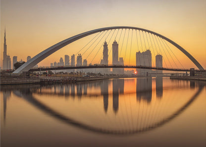 A panoramic photograph of the Dubai skyline at sunrise, viewed from across the water. A large, modern arched bridge spans the canal in the foreground, and its reflection creates a perfect oval shape in the calm water. In the background, skyscrapers, including the Burj Khalifa, are silhouetted against a glowing orange and yellow sky. The entire scene is presented within a silver picture frame. Wall Art