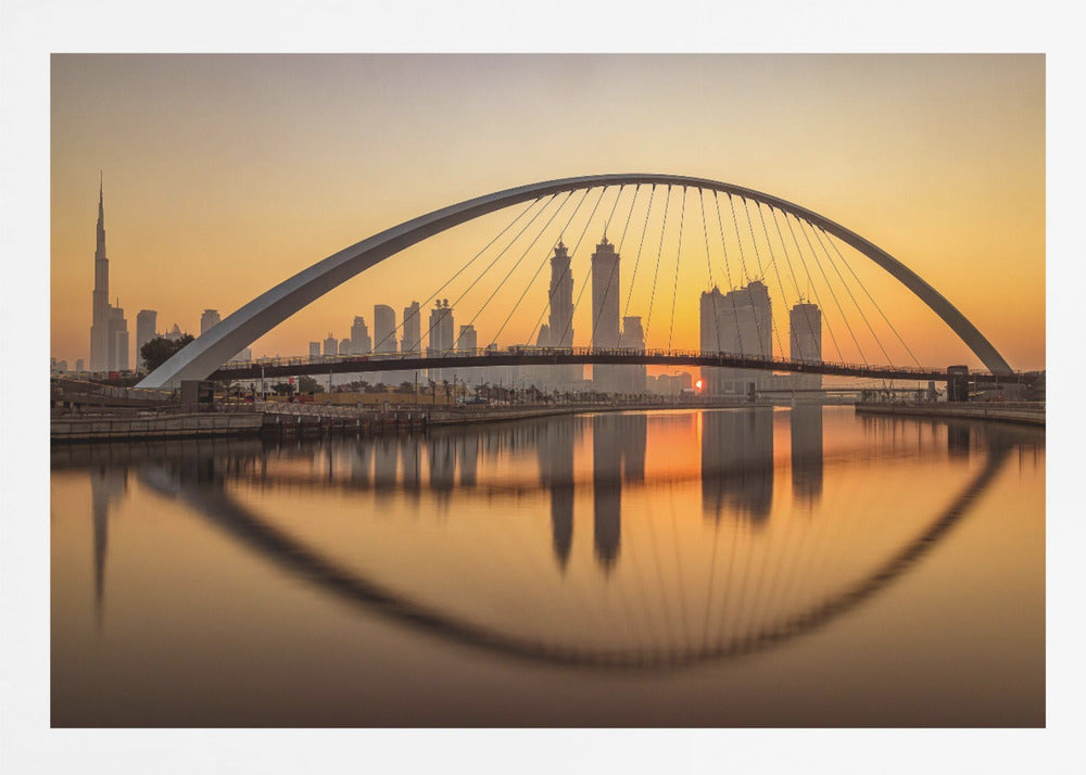 A panoramic photograph of the Dubai skyline at sunrise, viewed from across the water. A large, modern arched bridge spans the canal in the foreground, and its reflection creates a perfect oval shape in the calm water. In the background, skyscrapers, including the Burj Khalifa, are silhouetted against a glowing orange and yellow sky. The entire scene is presented within a silver picture frame. Wall Art
