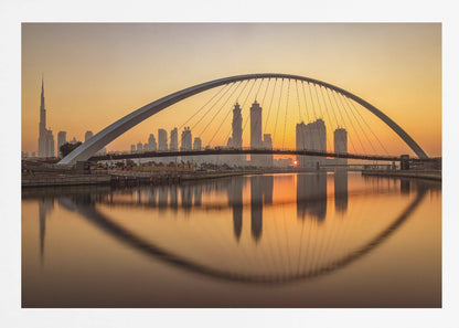 A panoramic photograph of the Dubai skyline at sunrise, viewed from across the water. A large, modern arched bridge spans the canal in the foreground, and its reflection creates a perfect oval shape in the calm water. In the background, skyscrapers, including the Burj Khalifa, are silhouetted against a glowing orange and yellow sky. The entire scene is presented within a silver picture frame. Wall Art