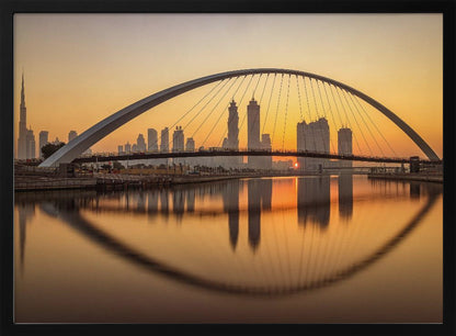 A panoramic photograph of the Dubai skyline at sunrise, viewed from across the water. A large, modern arched bridge spans the canal in the foreground, and its reflection creates a perfect oval shape in the calm water. In the background, skyscrapers, including the Burj Khalifa, are silhouetted against a glowing orange and yellow sky. The entire scene is presented within a silver picture frame. Wall Art