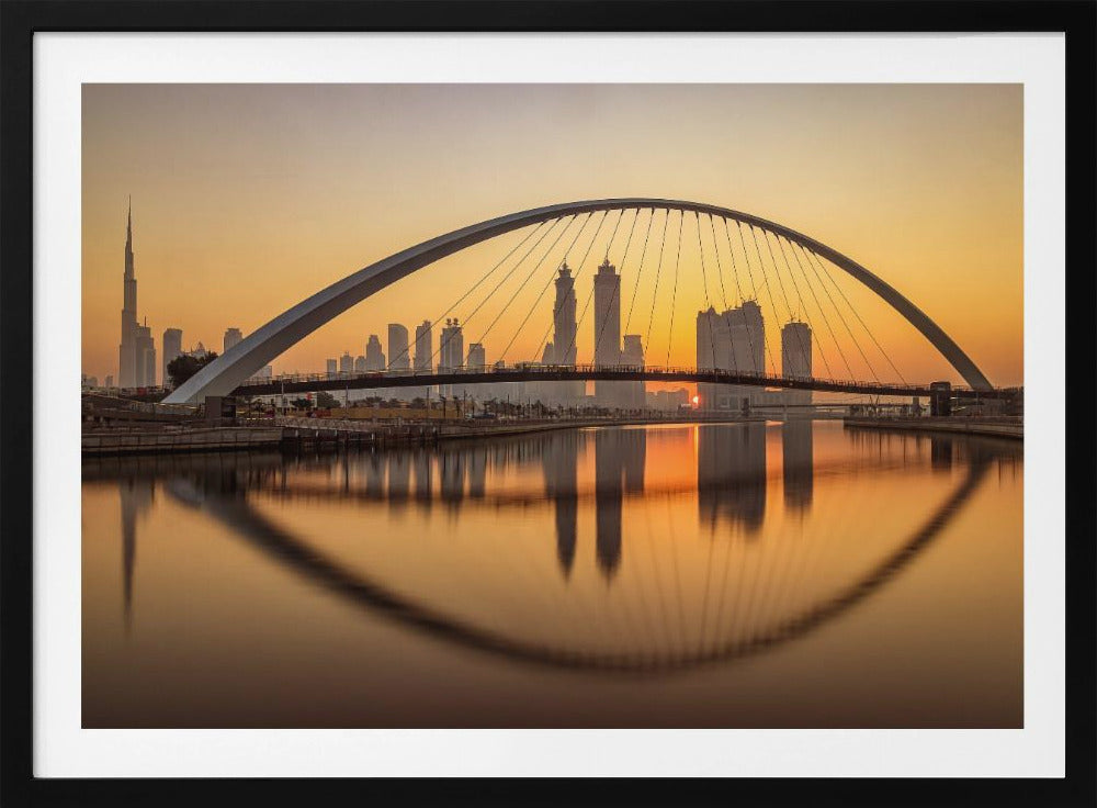A panoramic photograph of the Dubai skyline at sunrise, viewed from across the water. A large, modern arched bridge spans the canal in the foreground, and its reflection creates a perfect oval shape in the calm water. In the background, skyscrapers, including the Burj Khalifa, are silhouetted against a glowing orange and yellow sky. The entire scene is presented within a silver picture frame. Wall Art