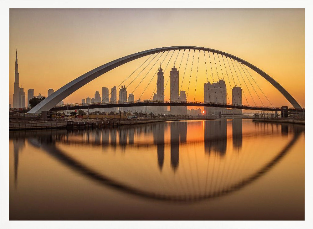 A panoramic photograph of the Dubai skyline at sunrise, viewed from across the water. A large, modern arched bridge spans the canal in the foreground, and its reflection creates a perfect oval shape in the calm water. In the background, skyscrapers, including the Burj Khalifa, are silhouetted against a glowing orange and yellow sky. The entire scene is presented within a silver picture frame. Wall Art
