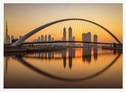 A panoramic photograph of the Dubai skyline at sunrise, viewed from across the water. A large, modern arched bridge spans the canal in the foreground, and its reflection creates a perfect oval shape in the calm water. In the background, skyscrapers, including the Burj Khalifa, are silhouetted against a glowing orange and yellow sky. The entire scene is presented within a silver picture frame. Wall Art