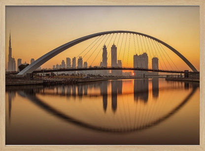 A panoramic photograph of the Dubai skyline at sunrise, viewed from across the water. A large, modern arched bridge spans the canal in the foreground, and its reflection creates a perfect oval shape in the calm water. In the background, skyscrapers, including the Burj Khalifa, are silhouetted against a glowing orange and yellow sky. The entire scene is presented within a silver picture frame. Wall Art