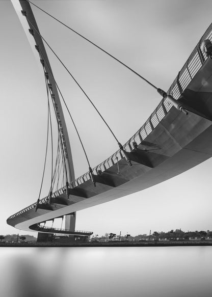 A dynamic low-angle, black and white photograph of a modern cable-stayed pedestrian bridge. The bridge curves elegantly over calm, reflective water, with its deck suspended by numerous cables attached to a tall, angled pylon, creating a striking graphic composition against a bright, clear sky. Decor