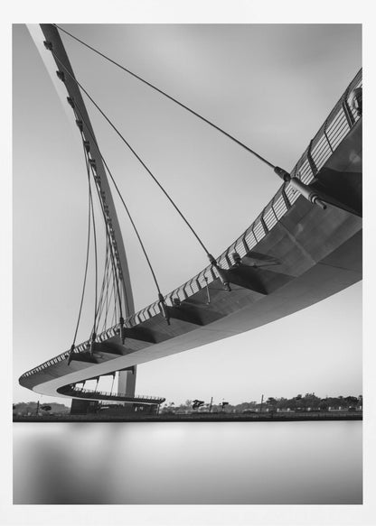 A dynamic low-angle, black and white photograph of a modern cable-stayed pedestrian bridge. The bridge curves elegantly over calm, reflective water, with its deck suspended by numerous cables attached to a tall, angled pylon, creating a striking graphic composition against a bright, clear sky. Decor